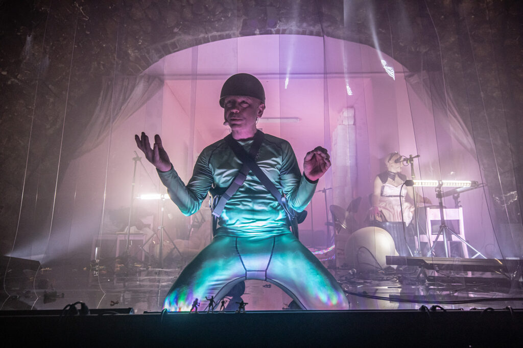 Zdjęcie ze spektaklu Saga rodu Szyców. Aktor w opalizującym kombinezonie, szelkach i hełmie klęczy i unosi dłonie. Przed nim stoją małe ludziki. Za kurtyną paskową z pleksi przy bębnach siedzi łysa aktorka z opaską z ćwiekami.