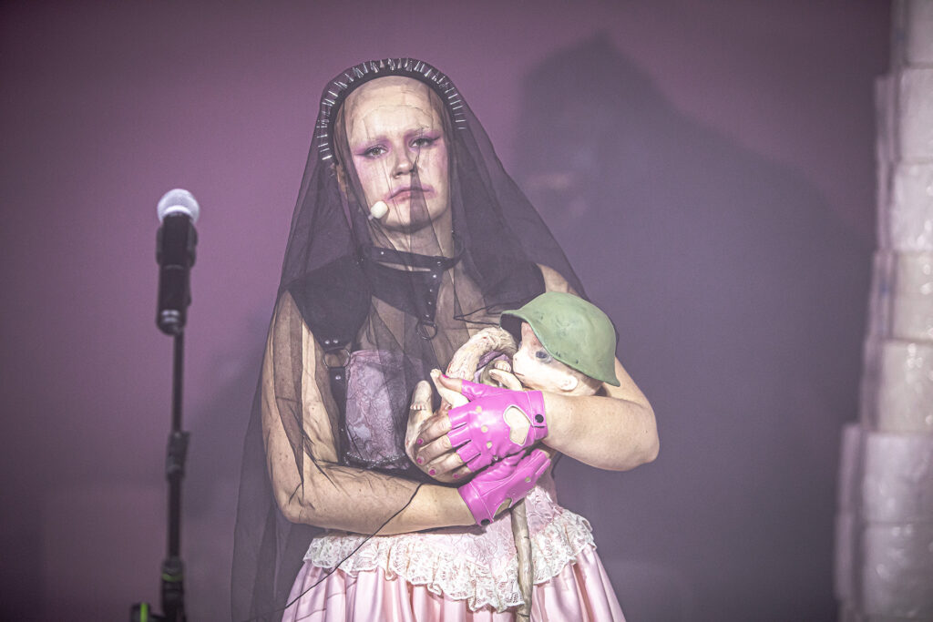 Zdjęcie ze spektaklu Saga rodu Szyców. Zbliżenie. Łysa aktorka ma opaskę z ćwiekami i czarny tiulowy welon. W ramionach trzyma lalkę noworodka w hełmie. Obok aktorki jest mikrofon. W tle jest figura goryla.