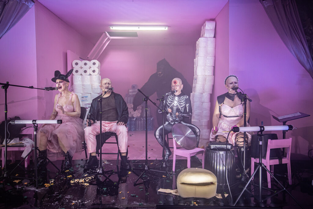 Zdjęcie ze spektaklu Saga rodu Szyców. Dwie aktorki i dwaj aktorzy siedzą przy instrumentach muzycznych i śpiewają do mikrofonów. Aktor ma strój kościotrupa i krew na czole. Pod ścianą stoją zapasy papieru toaletowego, butelek wody i figura goryla