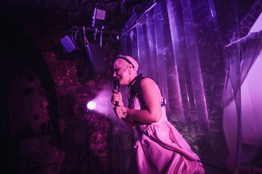 Zdjęcie ze spektaklu Saga rodu Szyców. Zdjęcie zabarwione na różowo. Łysa aktorka trzyma mikrofon na kablu. Aktorka ma sukienkę z gorsetem, skórzaną uprząż, skórzane rękawiczki, na głowie ma opaskę z ćwiekami. W tle jest kurtyna paskowa z pleksi.