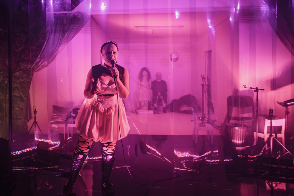 Zdjęcie ze spektaklu Saga rodu Szyców. Łysa aktorka trzyma mikrofon na kablu. Aktorka ma sukienkę z gorsetem, opaskę z ćwiekami skórzaną uprząż, rękawiczki, długie buty i potargane rajstopy. W tle za kurtyną z pleksi siedzą aktor i aktorka.