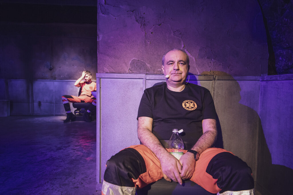 Zdjęcie ze spektaklu Pogo. Starszy aktor w stroju ratownika medycznego siedzi oparty o ścianę. Między nogami trzyma butelkę wody. W tle na krześle siedzi młody aktor w stroju ratownika medycznego i trzyma się za głowę