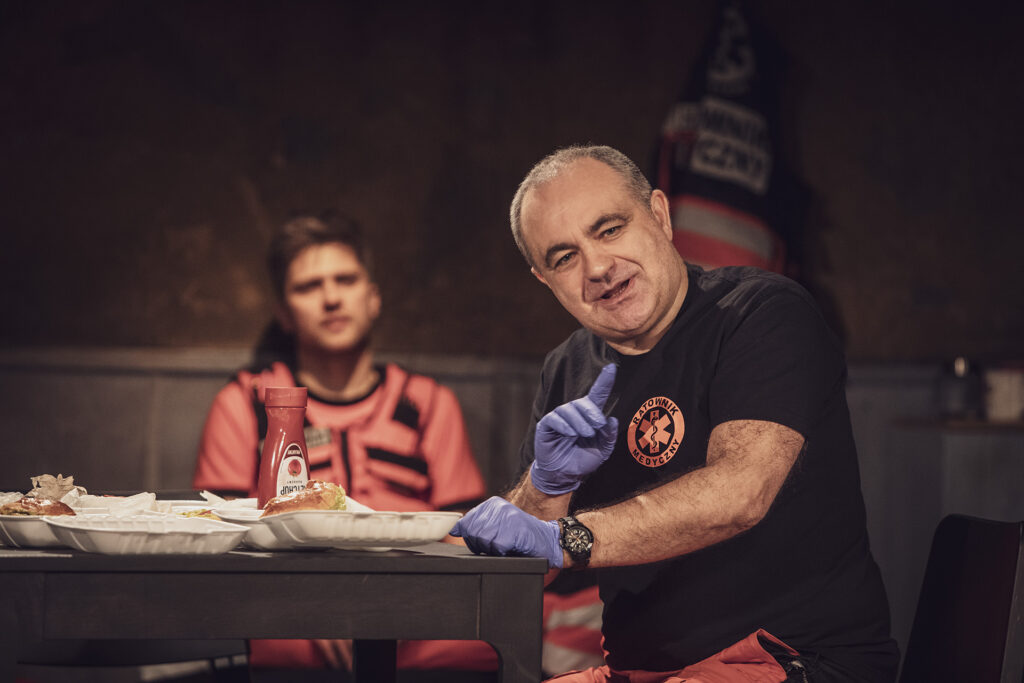Zdjęcie ze spektaklu Pogo. Starszy aktor w stroju ratownika medycznego siedzi przy stole. Ma rękawiczki, unosi jeden palec wskazujący. Na stole są hamburgery w opakowaniach na wynos i ketchup. W tle siedzi młody aktor w stroju ratownika medycznego.