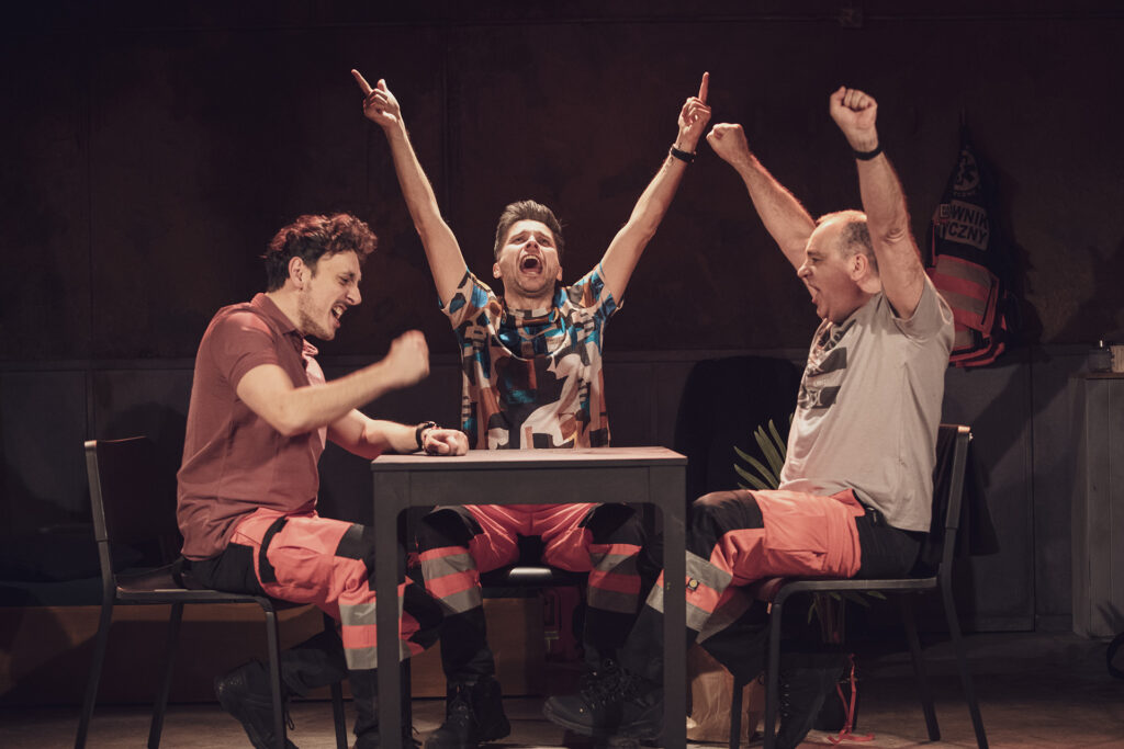 Zdjęcie ze spektaklu Pogo. Trzej aktorzy siedzą przy stole. Mają spodnie ratowników medycznych i codzienne koszulki. Wszyscy śpiewają. Dwaj unoszą ręce do góry, jeden bije pięścią w stół.