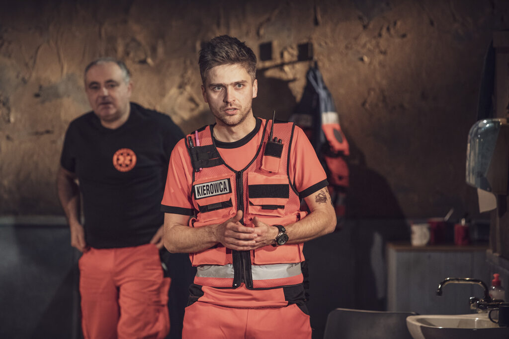 Zdjęcie ze spektaklu Pogo. Młody aktor w stroju ratownika medycznego z napisem kierowca. Trzyma dłonie przed sobą, ma poważną minę. W tle jest starszy aktor w stroju ratownika medycznego.