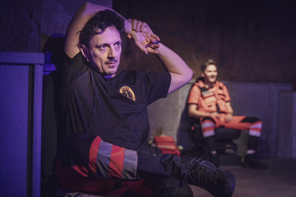 Zdjęcie ze spektaklu Pogo. Aktor w stroju ratownika medycznego trzyma ręce za głowę, jedną nogę opiera na kolanie. W tle siedzi drugi aktor w stroju ratownika medycznego.