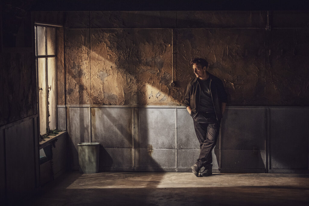 Zdjęcie ze spektaklu Pogo. Aktor w ciemnym stroju stoi oparty o ścianę, trzyma ręce w kieszeniach. Ściany i podłoga są ciemne. Po lewej stronie jest okno.