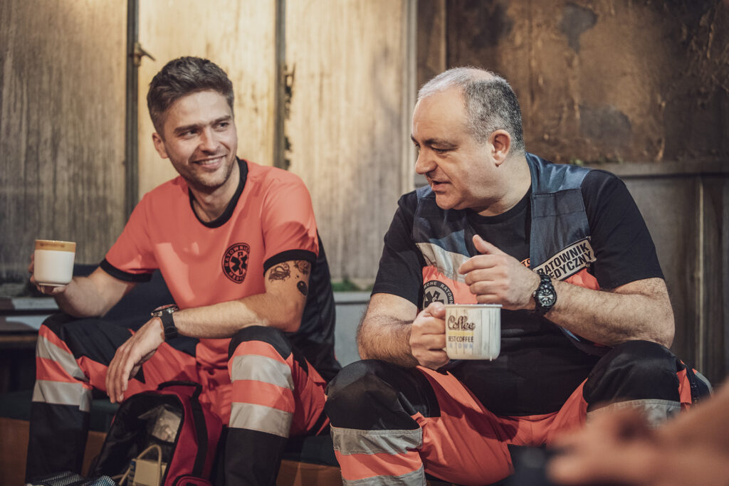 Zdjęcie ze spektaklu Pogo. Młodszy i starszy aktor w strojach ratowników medycznych siedzą obok siebie, patrzą na siebie i się uśmiechają. W dłoniach trzymają kubki. Młodszy aktor ma tatuaże na ręce.