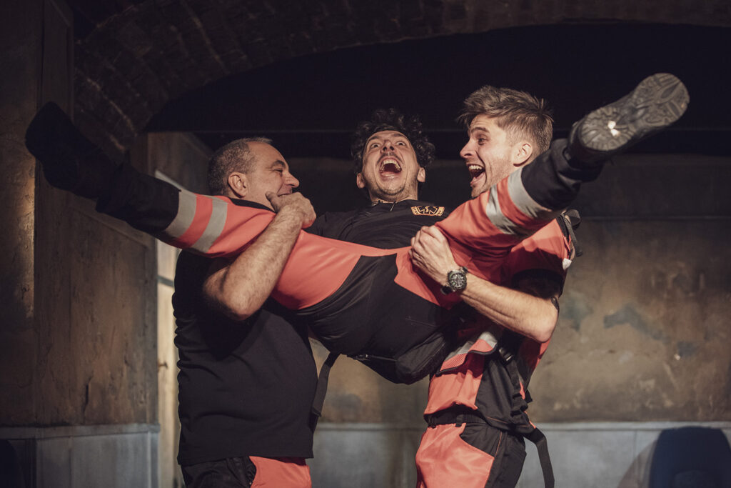 Zdjęcie ze spektaklu Pogo. Dwaj aktorzy trzymają trzeciego aktora za nogi i barki jakby go huśtali. Wszyscy się śmieją. Wszyscy są w strojach ratowników medycznych.