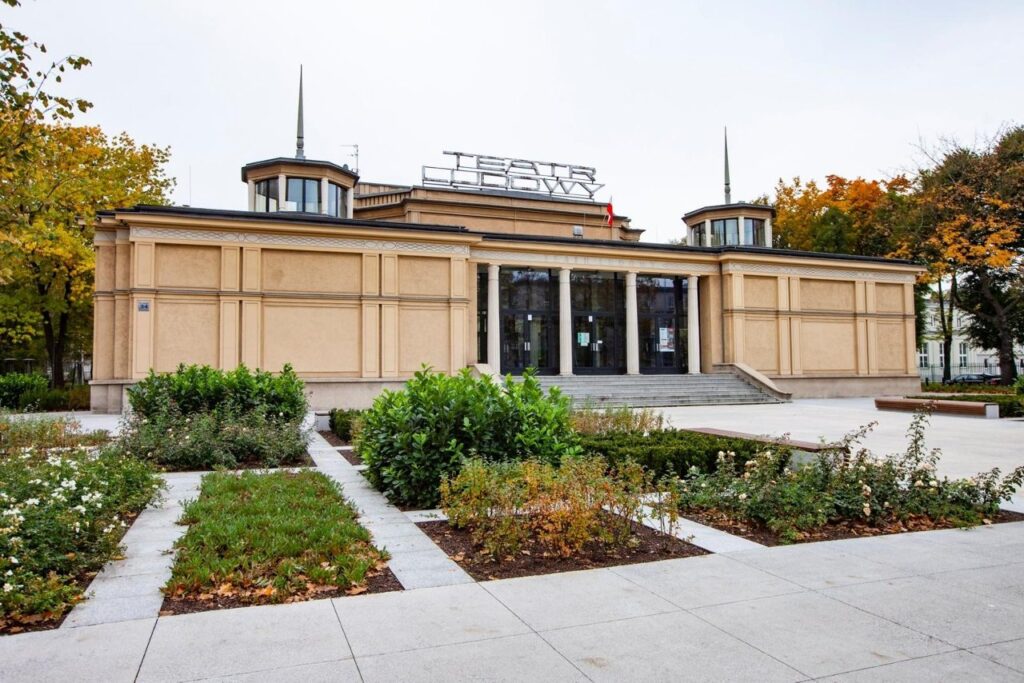 Zdjęcie budynku głównego Teatru Ludowego. Na pierwszym planie mamy chodnik i wygrodzone zielone trawniki. Na drugim planie jest budynek. Przeszklone wejście znajduje się po środku budynku. Po bokach na dachu widać dwa świetliki - wieżyczki, po środku jest duży neon z napisem Teatr Ludowy.