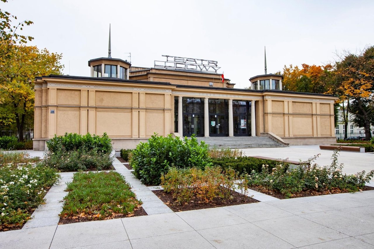 Zdjęcie budynku głównego Teatru Ludowego. Na pierwszym planie mamy chodnik i wygrodzone zielone trawniki. Na drugim planie jest budynek. Przeszklone wejście znajduje się po środku budynku. Po bokach na dachu widać dwa świetliki - wieżyczki, po środku jest duży neon z napisem Teatr Ludowy.