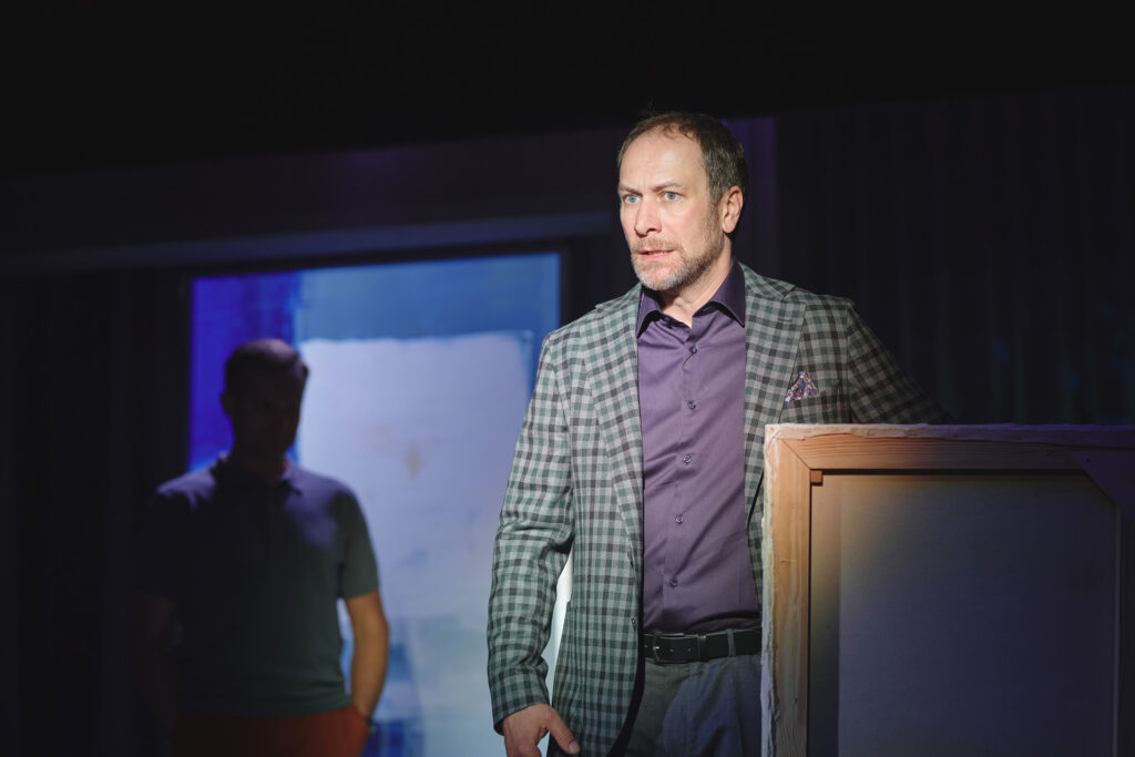 Zdjęcie ze spektaklu Sztuka. Zbliżenie. Aktor w marynarce stoi przed obrazem. W tle stoi aktor w polówce. Za nim na ścianie jest obraz przedstawiający kwadrat na niebieskim tle.