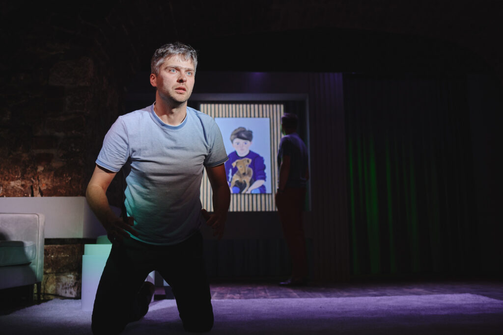 Zdjęcie ze spektaklu Sztuka. Aktor w koszulce z krótkim rękawem klęczy. W tle na ścianie jest portret małego chłopca z psem. Przy obrazie stoi drugi aktor.