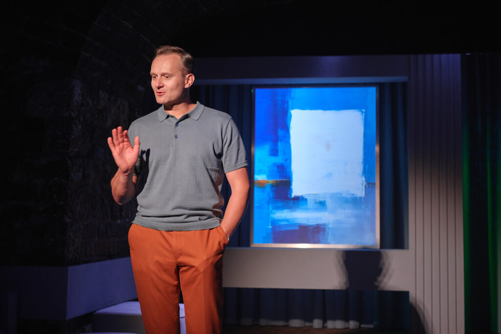 djęcie ze spektaklu Sztuka. Aktor w polówce stoi na środku. Jedną rękę trzyma w kieszeni, a drugą unosi. Za nim jest obraz przedstawiający kwadrat na niebieskim tle.