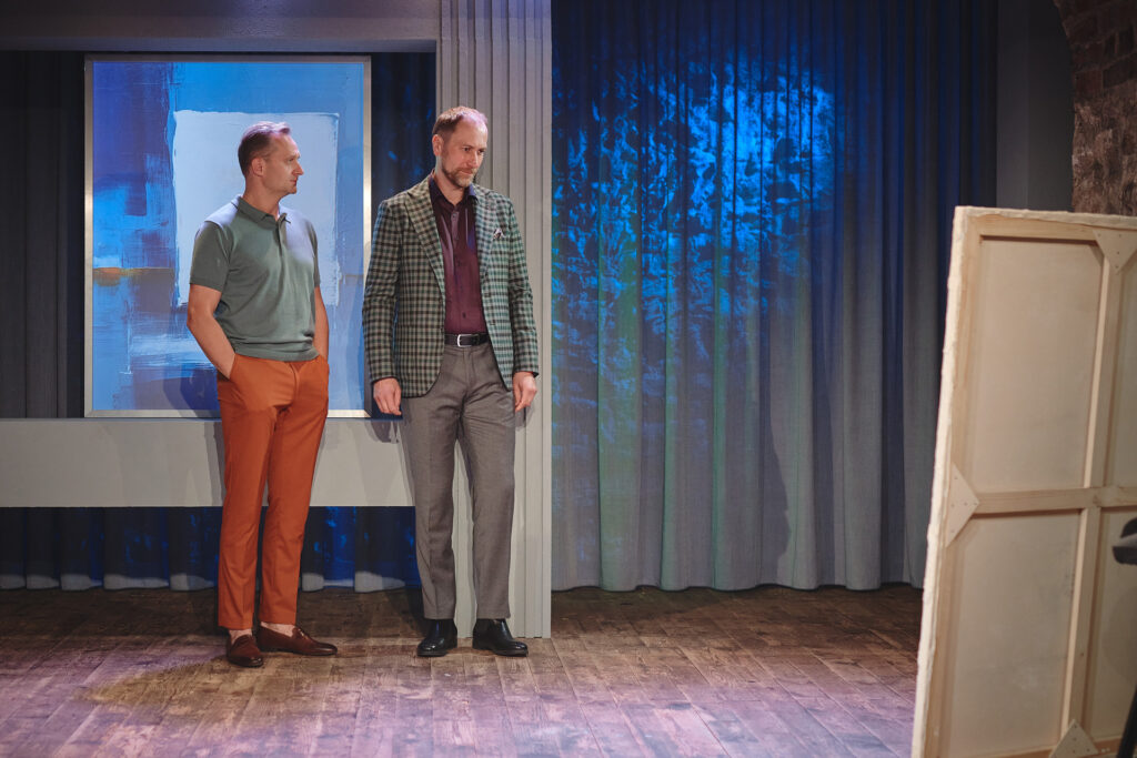 Zdjęcie ze spektaklu Sztuka. Dwaj aktorzy przed płótnem. Aktor w polówce trzyma ręce w kieszeniach i patrzy na aktora w marynarce. Za nimi jest obraz przedstawiający kwadrat na niebieskim tle. Z tyłu jest zasłona podświetlona na niebiesko.