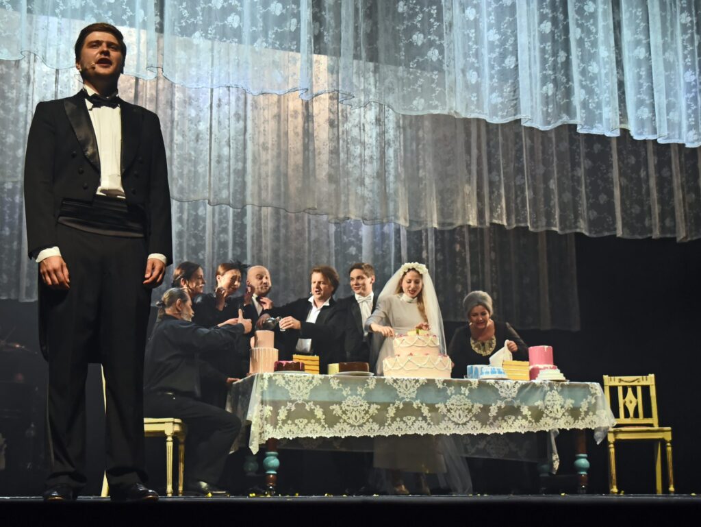 djęcie ze spektaklu Zahipnotyzuj mnie. Na brzegu sceny stoi aktor w smokingu. Za nim przy stole para młoda kroi tort. Wokół nich są pozostali aktorzy i aktorki. Na stole stoi jeszcze kilka małych tortów. Nad sceną zwisają koronkowe zasłony.