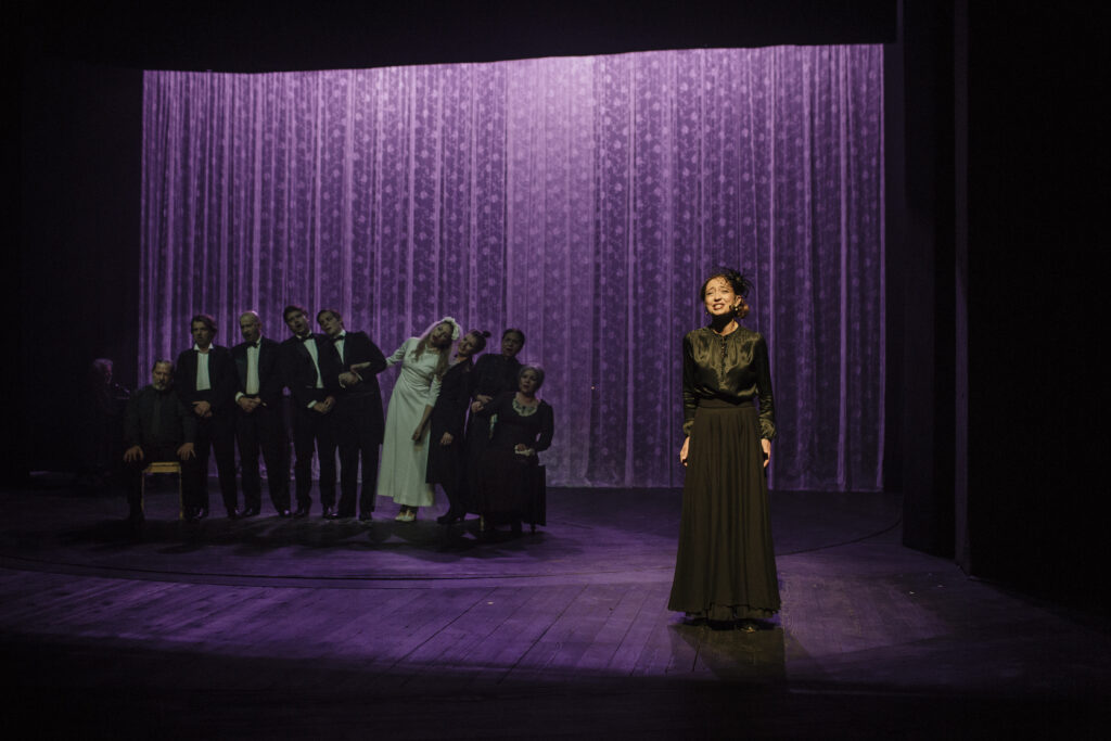 Zdjęcie ze spektaklu Zahipnotyzuj mnie. Aktorka w ciemnej długiej sukni z woalką na głowie. W tle aktorzy w garniturach, aktorki w czarnych sukniach i jedna aktorka w sukni ślubnej. W tle na ścianie koronkowe zasłony podświetlone na fioletowo.