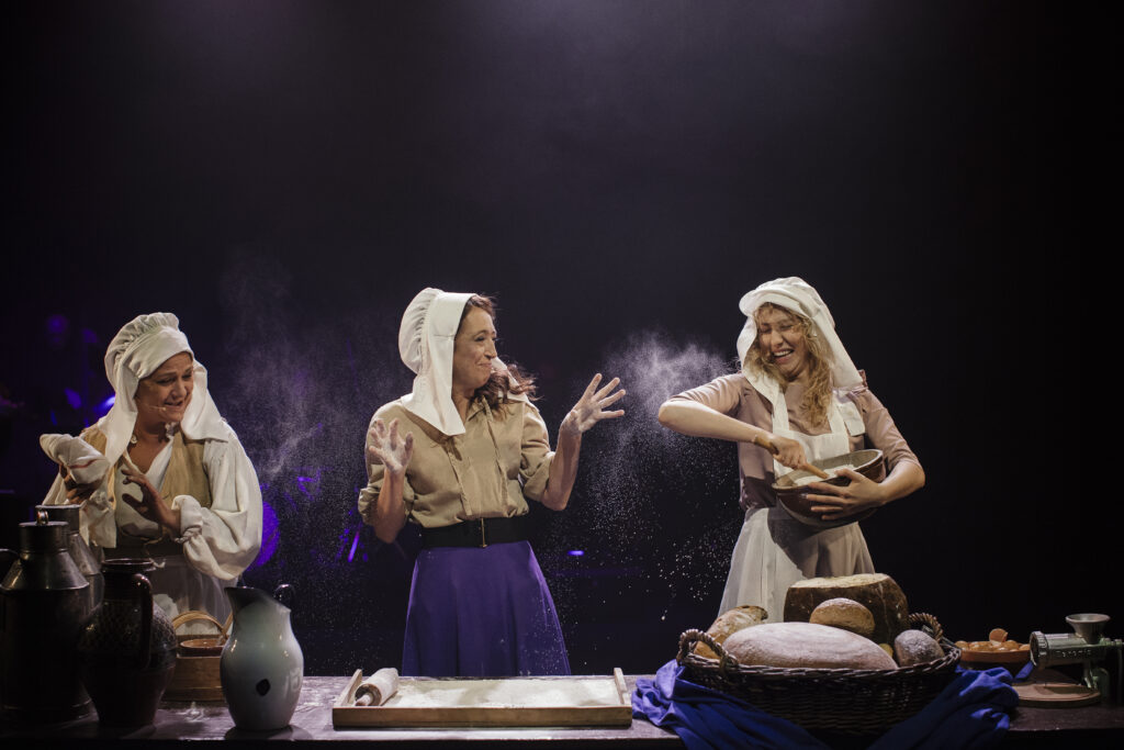 Zdjęcie ze spektaklu Zahipnotyzuj mnie. Trzy aktorki w czepkach na głowie stoją przy stole. Na stole jest stolnica z wałkiem, mąka i koszyk pieczywa. Jedna aktorka miesza coś w garnku. Druga rozsypuje w powietrzu mąkę.