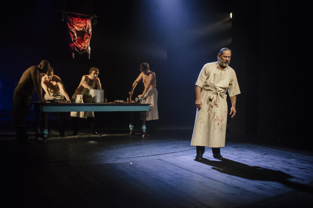 Zdjęcie ze spektaklu Zahipnotyzuj mnie. Starszy aktor z zarostem stoi w zakrwawionym fartuchu. W tle czterej półnadzy aktorzy w fartuchach pracują przy stole z garnkami i maszynkami do mięsa. Nad stołem wisi materiał jak wypatroszone mięso.