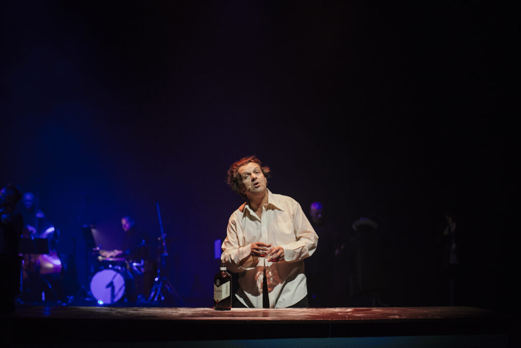 Zdjęcie ze spektaklu Zahipnotyzuj mnie. Aktor w koszuli stoi przy stole. Trzyma pustką szklankę. Na stole stoi butelka whisky. W tle aktorzy grają na instrumentach muzycznych.