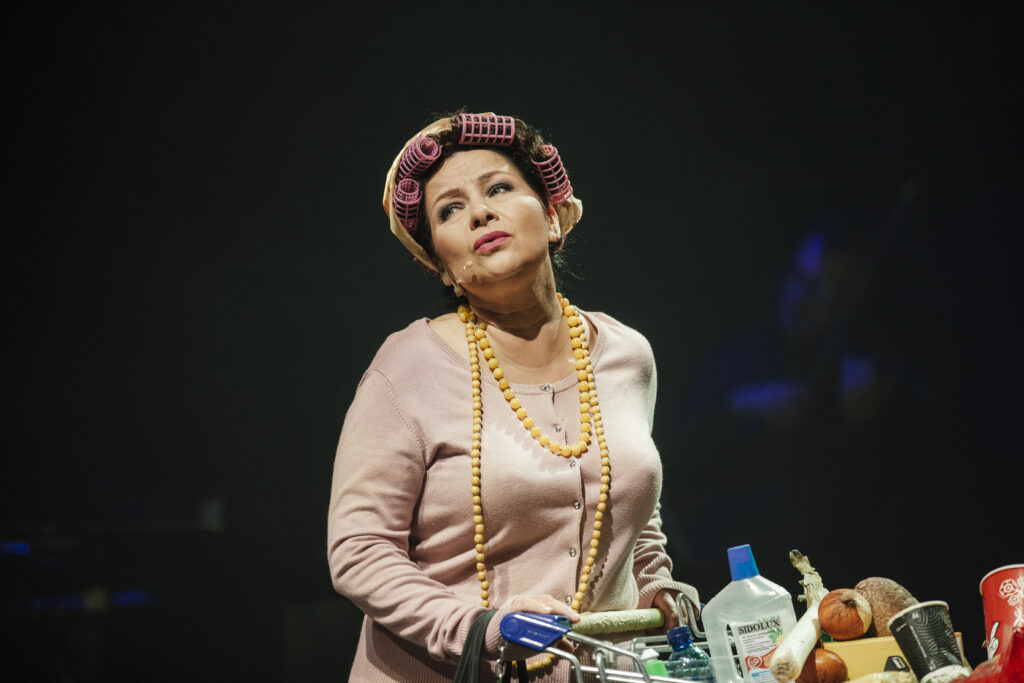 Zdjęcie ze spektaklu Zahipnotyzuj mnie. Zbliżenie. Aktorka w koralach i wałkach na głowie pcha wózek sklepowy wypełniony jedzeniem i produktami higienicznymi.