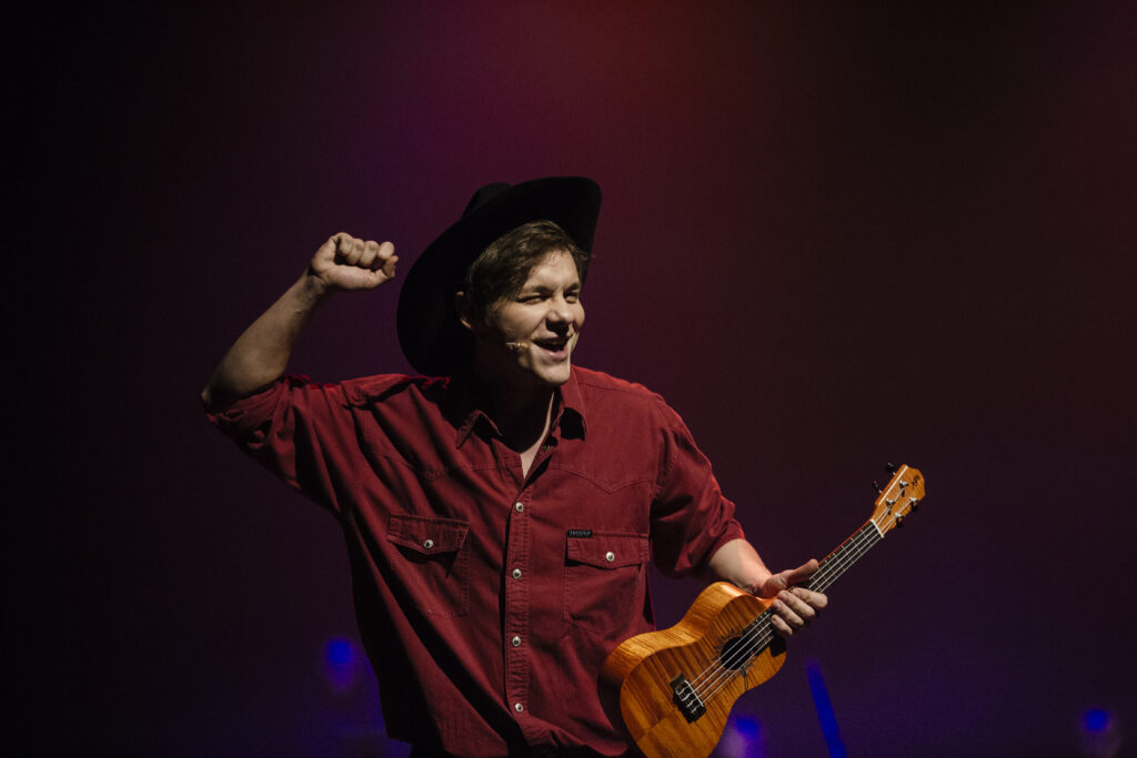 Zdjęcie ze spektaklu Zahipnotyzuj mnie. Zbliżenie. Aktor w koszuli i kowbojskim kapeluszu śpiewa. W jednej ręce trzyma ukulele. Drugą rękę zaciska w pięść i unosi.