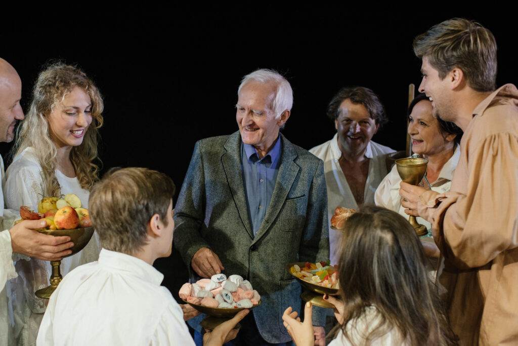 Zdjęcie ze spektaklu Zahipnotyzuj mnie Piosenki Zygmunta Koniecznego. Scena zbiorowa. W środku stoi Zygmunt Konieczny. Wokół aktorzy i aktorki częstują go owocami i słodyczami.