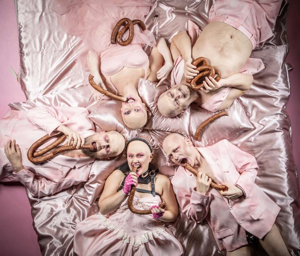 Zdjęcie ze spektaklu Saga rodu Szyców. Na różowym satynowym materiale leżą dwie aktorki i trzech aktorów. Wszyscy są łysi. Wszyscy mają różowe stroje. Wszyscy trzymają lub gryzą pęta kiełbasy.