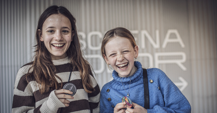 Dwie dziewczynki w wieku szkolnym. Uśnmiechnięte pozują do zdjęcia w rękach trzymają przypinki festiwalowe. W tle napis Scena Kolorowa.