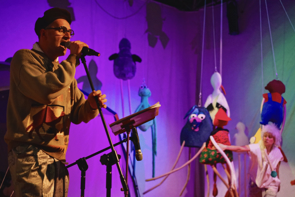 Zdjęcie ze spektaklu Lokomotywą przez świat. Aktor śpiewa do mikrofonu na statywie. Aktor ma sweter, spodnie z napisami, czapkę i okulary. W tle aktorka w peruce, z uszami elfa stoi między materiałowymi lalkami zwisającymi z sufitu.