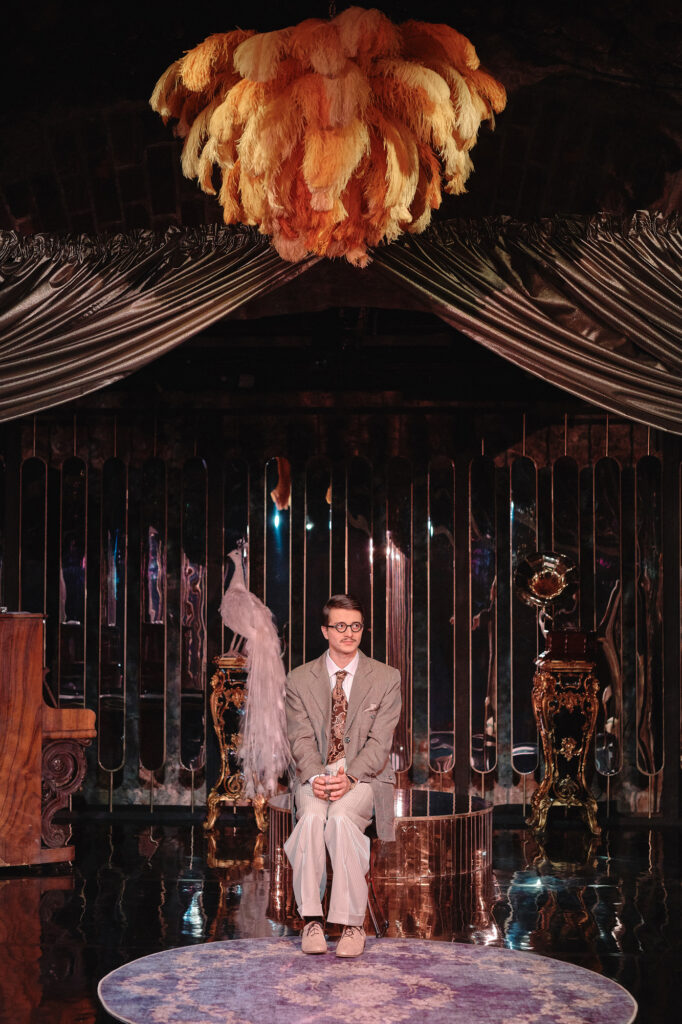 Zdjęcie ze spektaklu Boska. Młody aktor w garniturze i okularach siedzi na środku sceny. Scenografia przypomina wystrój z lat 20. Jest przepych, stoi figurka pawia, z sufitu zwisają pawie pióra.