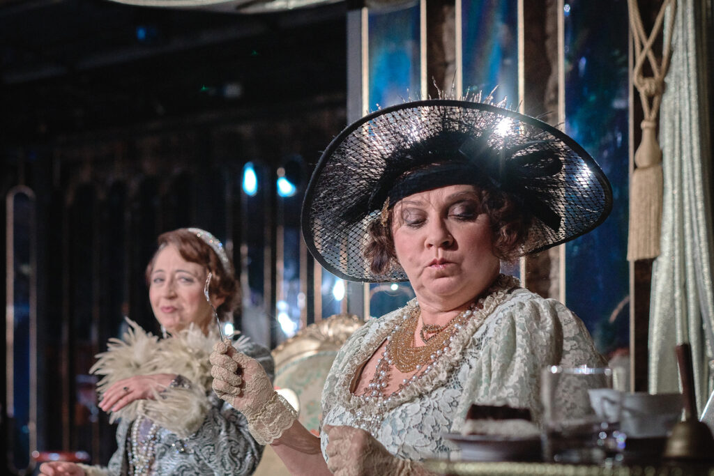 Zdjęcie ze spektaklu Boska. Zbliżenie. Aktorka w kapeluszu i rękawiczkach siedzi przy stole, spuszcza wzrok, w ręce trzyma łyżeczkę. Za nią siedzi druga aktorka i się uśmiecha