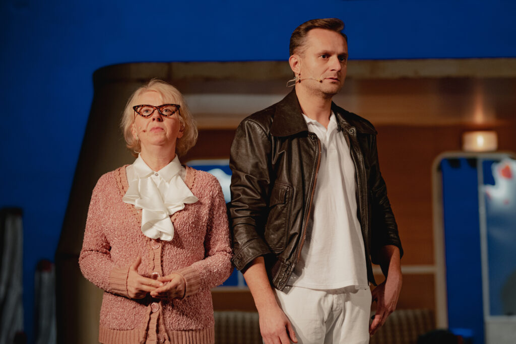 Zdjęcie ze spektaklu Don Kichot. Starsza aktorka w okularach, swetrze i koszuli z falbanką stoi obok młodego aktora w skórzanej kurtce.