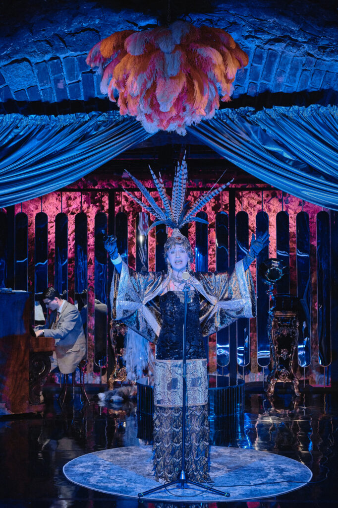 Zdjęcie ze spektaklu Boska. Aktorka stoi na scenie i śpiewa do mikrofonu na statywie. Ma długą błyszczącą suknię, na głowie ma pióropusz z piór bażanta. Z sufitu zwisają pawie pióra. W tle mężczyzna w garniturze siedzi przy pianinie.