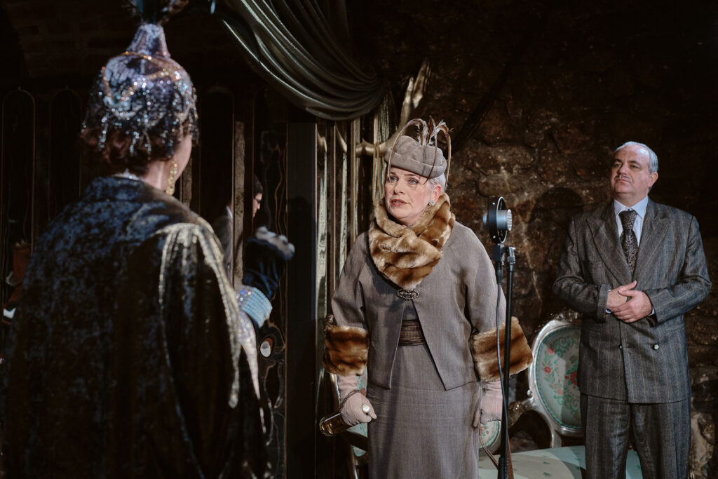Zdjęcie ze spektaklu Boska. Dwie aktorki patrzą na siebie. Aktorka w pióropuszu stoi tyłem. Druga aktorka ma garsonkę ozdobioną futrem, na głowie ma toczek, w ręce trzyma butelkę wina. Z tyłu stoi starszy aktor w garniturze i patrzy na aktorki.