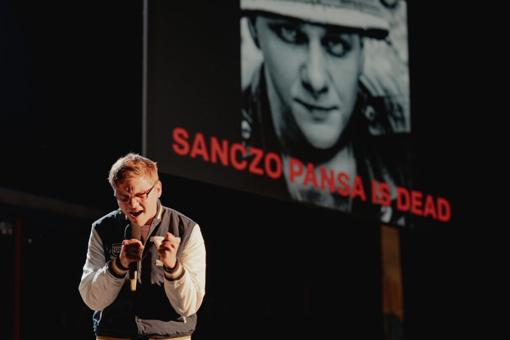 djęcie ze spektaklu Don Kichot. Aktor w bejsbolówce i okularach krzyczy do mikrofonu. Za nim na ścianie jest wyświetlone jego zdjęcie z podpisem Sanczo Pansa is dead.