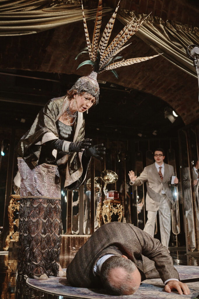 Zdjęcie ze spektaklu Boska. Na podłodze leży aktor. Nad nim pochyla się aktorka w długiej sukni i pióropuszu z piór bażanta. W tle stoi mężczyzna w okularach i garniturze.