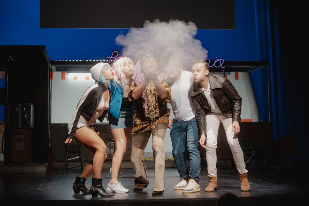 Zdjęcie ze spektaklu Don Kichot. Zdjęcie grupowe. Aktorzy w kolorowych strojach stoją w grupie i dmuchają przed siebie, powstaje przed nimi obłok szarego pyłu.