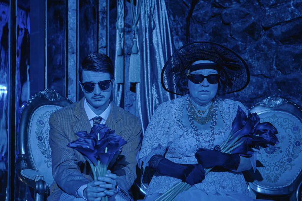 Zdjęcie ze spektaklu Boska. Zdjęcie zabarwione na niebiesko. Na fotelach obok siebie siedzą aktorka w kapeluszu i rękawiczkach i młody aktor w garniturze. Obydwoje mają okulary przeciwsłoneczne. W rękach trzymają bukiety kalii.