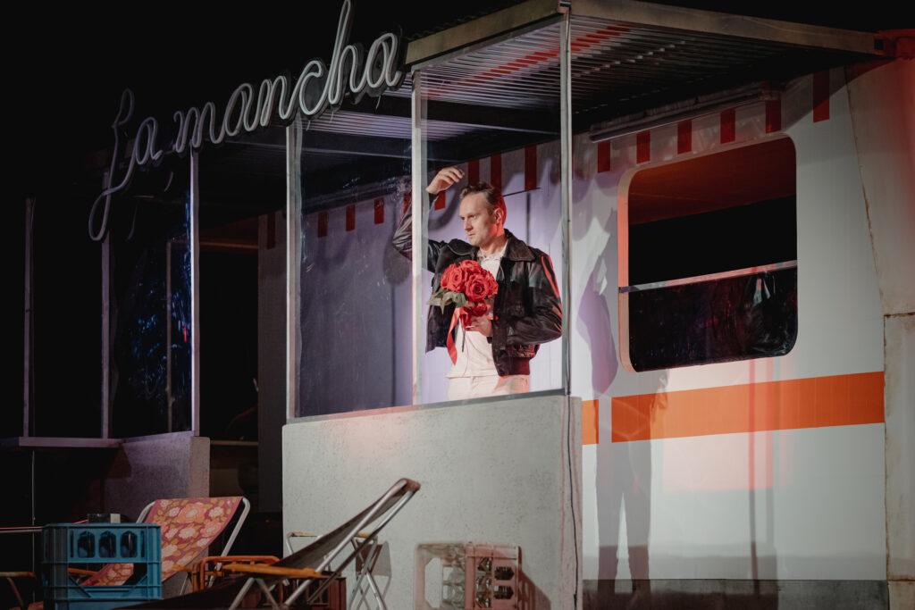 Zdjęcie ze spektaklu Don Kichot. Aktor w skórzanej kurtce trzyma w ręce bukiet róż. Stoi na werandzie kampera z napisem La Mancha. Przed kamperem są leżaki i skrzynki po napojach.