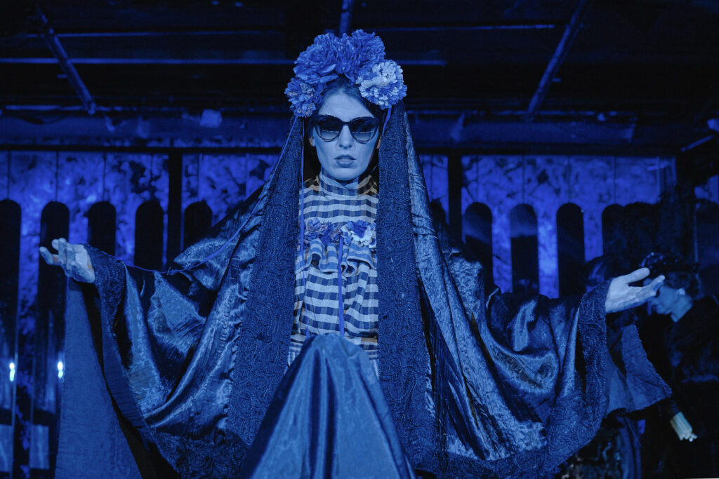 Zdjęcie ze spektaklu Boska. Zdjęcie zabarwione na niebiesko. Młoda aktorka stoi przodem i podnosi ręce. Ma na sobie suknię w kratę, czarną pelerynę, ma okulary przeciwsłoneczne i wianek na głowie.