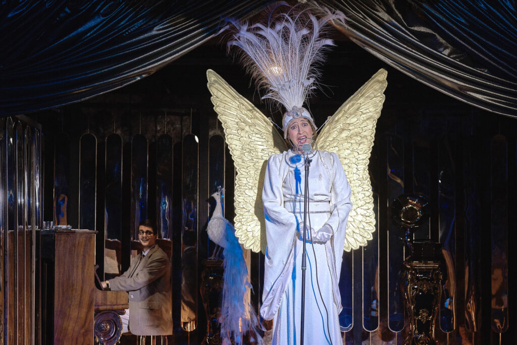 Zdjęcie ze spektaklu Boska. Aktorka stoi na scenie i śpiewa do mikrofonu na statywie. Ma długą suknię, na plecach ma duże skrzydła, a na głowie pióropusz z pawich piór. Za nią jest figurka pawia. W tle mężczyzna w garniturze siedzi przy pianinie.