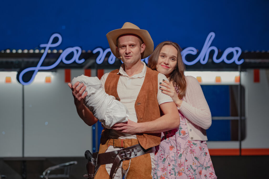 Zdjęcie ze spektaklu Don Kichot. Młody aktor w stroju kowboja trzyma w ręce zawiniątko z noworodkiem. Obejmuje go młoda aktorka. Za nimi jest kamper z napisem La Mancha.