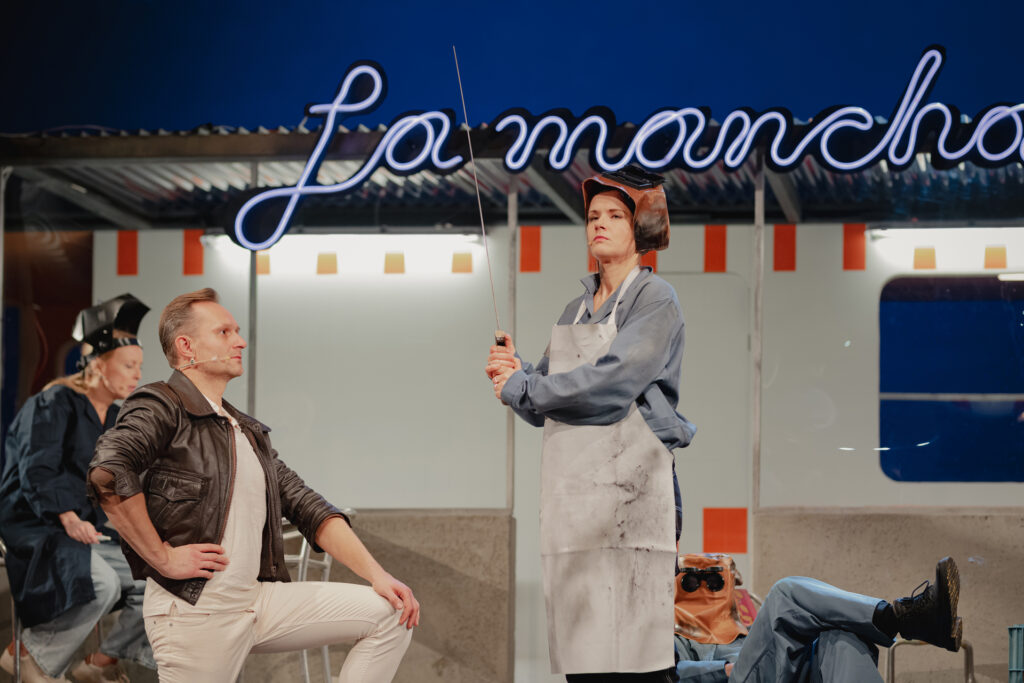 Zdjęcie ze spektaklu Don Kichot. Aktor w skórzanej kurtce klęka na jedno kolano. Przed nim stoi aktorka w stroju hutnika ze szpadą. Scena wygląda jak mianowanie na rycerza. Z tyłu siedzi aktorka i aktor. Za nimi jest kamper z napisem La Mancha.