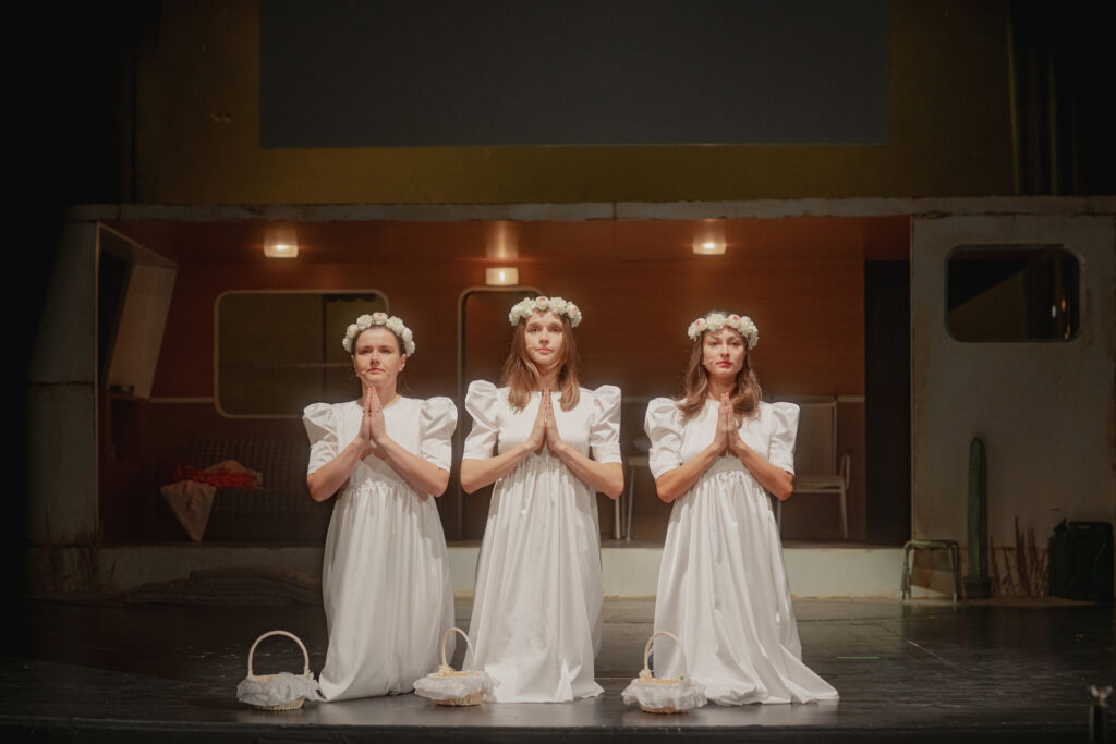 Zdjęcie ze spektaklu Don Kichot. Trzy młode aktorki klęczą obok siebie, mają ręce złożone jak do modlitwy. Aktorki mają długie sukienki, wianki na głowach, przed nimi są koszyczki do sypania kwiatów.