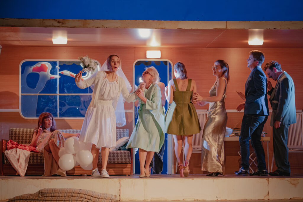 Zdjęcie ze spektaklu Don Kichot. Scena zbiorowa. Aktorki i aktorzy mają eleganckie stroje imprezowe. Na środku jest aktorka w białej sukni, w welonie i z kwiatami w dłoni. Z tyłu jest wnętrze kampera.