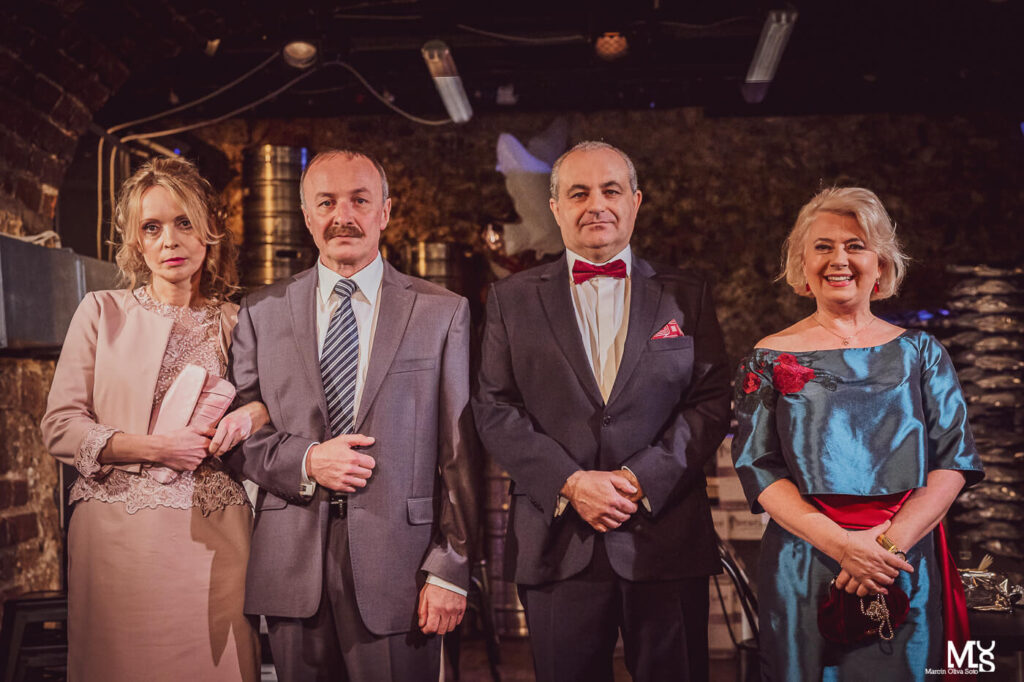 Zdjęcie ze spektaklu Wstyd. Dwie pary aktorskie, wszyscy stoją obok siebie w rządku. Są elegancko ubrani, panowie trzymają pod rękę Panie.
