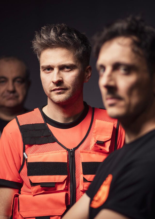 Zdjęcie z sesji wizerunkowej do spektaklu Pogo. Trzech mężczyzn ustawionych w lini jeden za drugim. Ubrani w stroje ratowników medycznych. Wyostrzenie obrazu na środkowego.