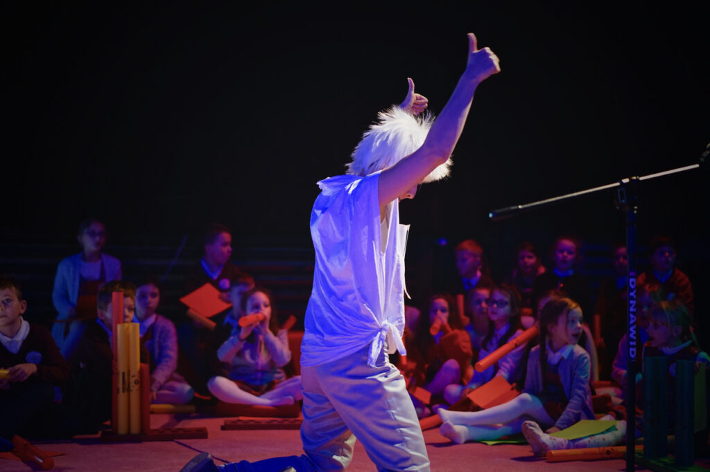 Zdjęcie ze spektaklu Lokomotywą przez świat. Aktorka w peruce stoi bokiem i unosi dwa kciuki w górę. W tle na scenie siedzą dzieci.