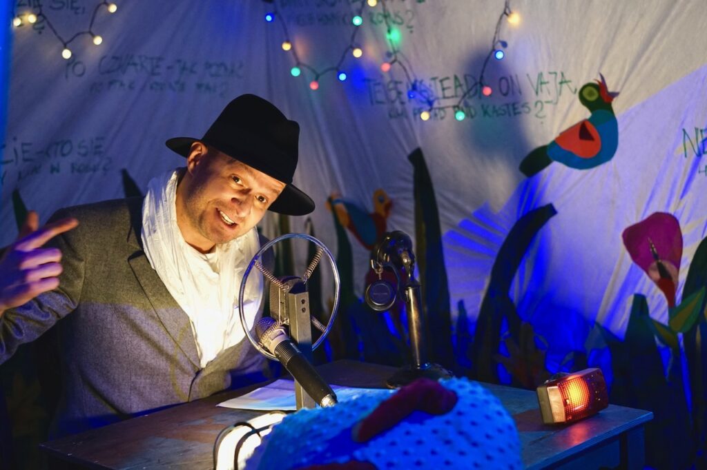 Zdjęcie ze spektaklu Lokomotywą przez świat. Zbliżenie. Aktor w kapeluszu i szalu siedzi przy biurku. Na biurku stoi mikrofon i słuchawki. Na ścianach są kolorowe lampki, rysunki ptaków i napisy.